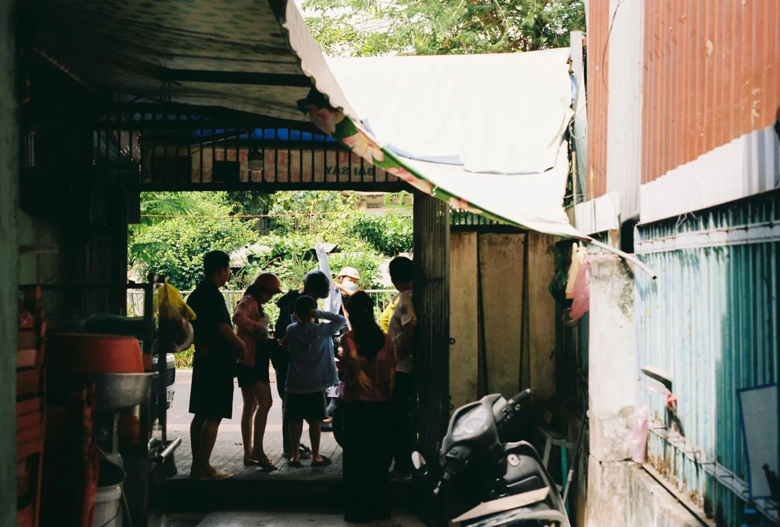 People gathered at the sunlit end of a covered alley, a motorbike parked to the side